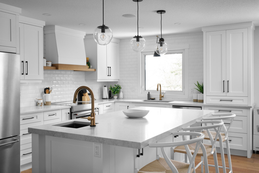Example of a transitional l-shaped medium tone wood floor and brown floor kitchen design with an undermount sink, shaker cabinets, white cabinets, white backsplash, subway tile backsplash, stainless steel appliances, an island and white countertops