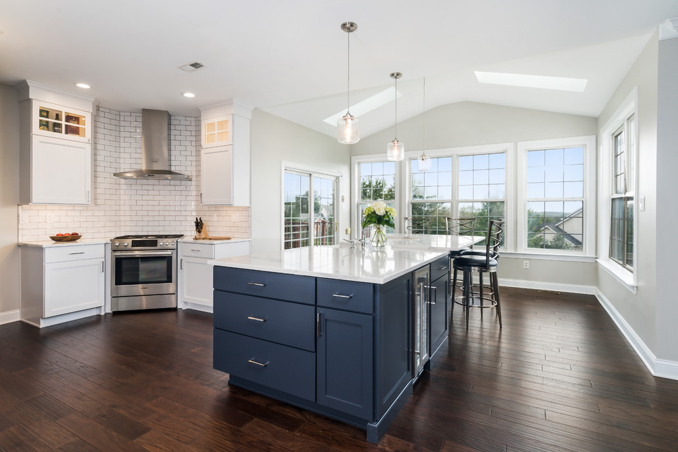 This is an example of a large transitional l-shaped eat-in kitchen in Philadelphia with a farmhouse sink, shaker cabinets, white cabinets, quartz benchtops, white splashback, subway tile splashback, stainless steel appliances, dark hardwood floors, with island, brown floor and white benchtop.