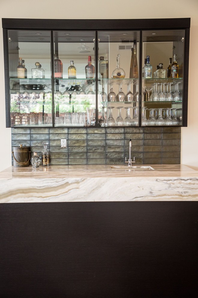 This is an example of a contemporary galley wet bar in Los Angeles with a drop-in sink, glass-front cabinets, black cabinets, onyx benchtops, grey splashback, glass tile splashback, medium hardwood floors and brown floor.