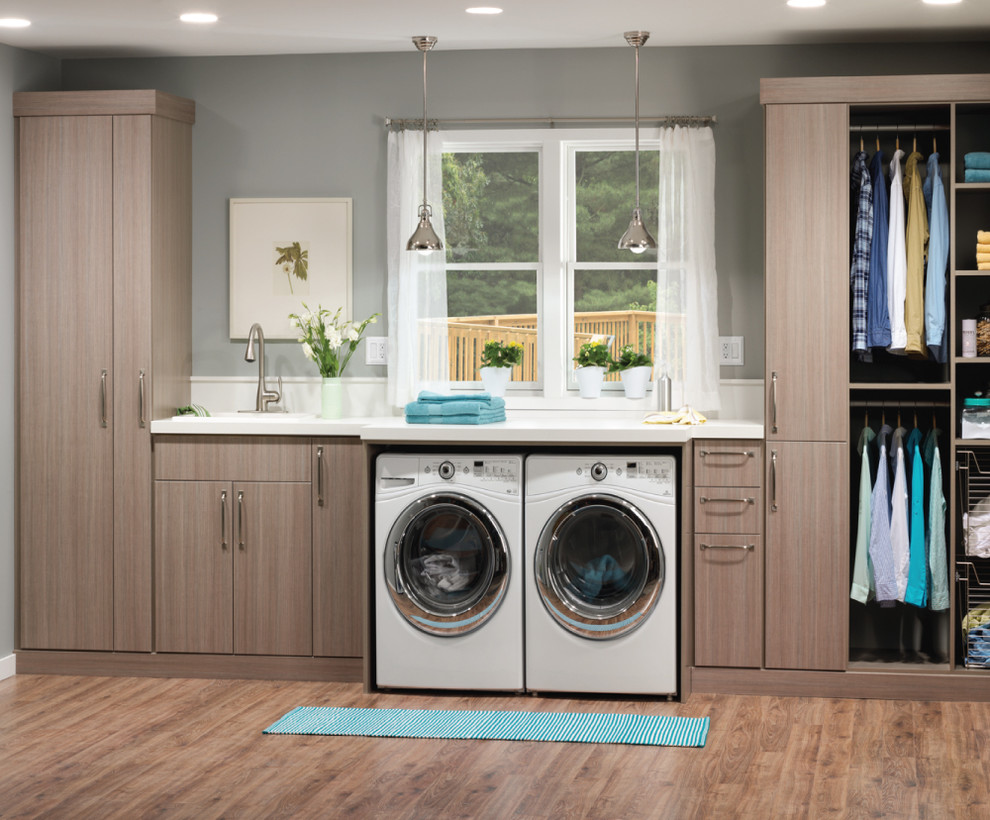 Inspiration for a mid-sized industrial single-wall dedicated laundry room in New York with a drop-in sink, flat-panel cabinets, a side-by-side washer and dryer, grey walls, light hardwood floors and grey cabinets.