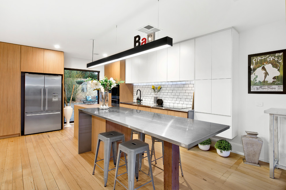 Mid-sized contemporary l-shaped open plan kitchen in Melbourne with an undermount sink, flat-panel cabinets, white cabinets, stainless steel benchtops, white splashback, subway tile splashback, stainless steel appliances, with island, grey benchtop, light hardwood floors and beige floor.