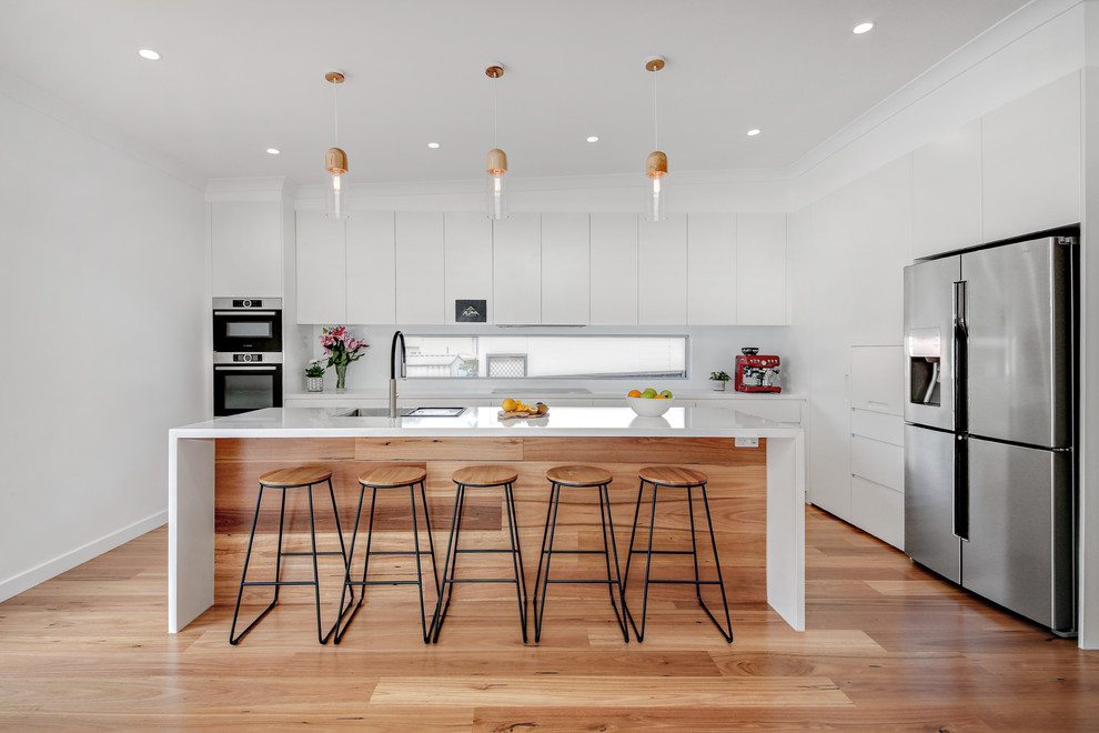 This is an example of a large modern l-shaped kitchen in Sydney with an undermount sink, flat-panel cabinets, white cabinets, quartz benchtops, white splashback, glass sheet splashback, stainless steel appliances, with island, brown floor, white benchtop and medium hardwood floors.