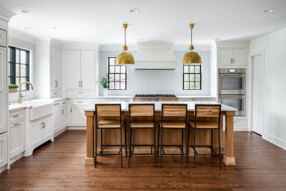 Design ideas for an expansive transitional l-shaped eat-in kitchen in New York with a farmhouse sink, white cabinets, quartz benchtops, white splashback, engineered quartz splashback, medium hardwood floors, with island, brown floor, white benchtop, shaker cabinets and stainless steel appliances.