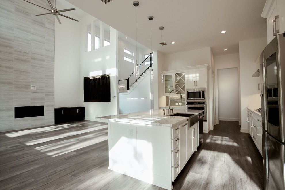Photo of a large modern l-shaped open plan kitchen in Dallas with a farmhouse sink, recessed-panel cabinets, white cabinets, granite benchtops, grey splashback, glass tile splashback, stainless steel appliances, medium hardwood floors, with island, brown floor and white benchtop.