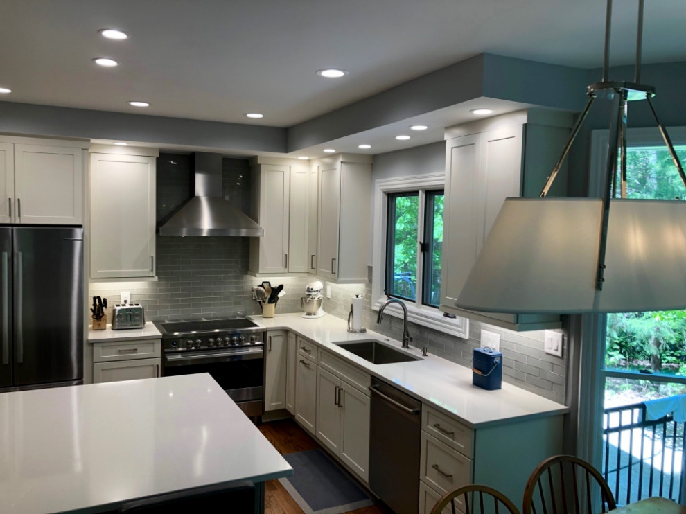 Large classic l-shaped kitchen/diner in Raleigh with a submerged sink, shaker cabinets, white cabinets, engineered stone countertops, grey splashback, glass tiled splashback, stainless steel appliances, dark hardwood flooring, an island, brown floors and white worktops.