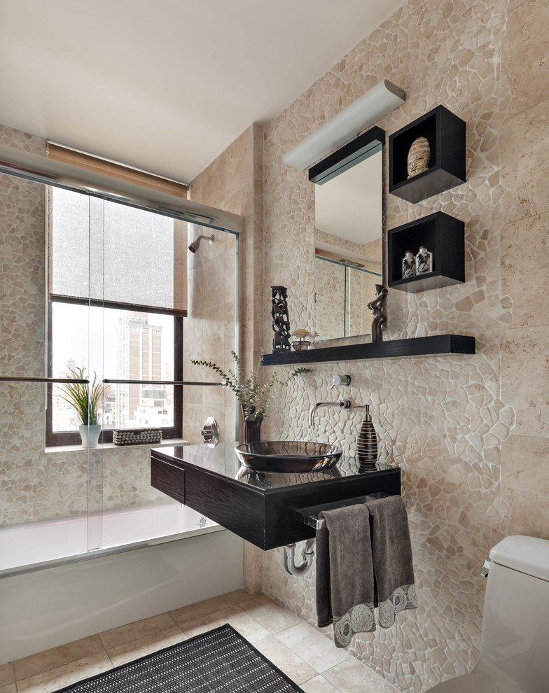 Inspiration for an eclectic 3/4 bathroom in New York with flat-panel cabinets, black cabinets, an alcove tub, a shower/bathtub combo, a one-piece toilet, beige tile, beige walls, a vessel sink, beige floor and a sliding shower screen.