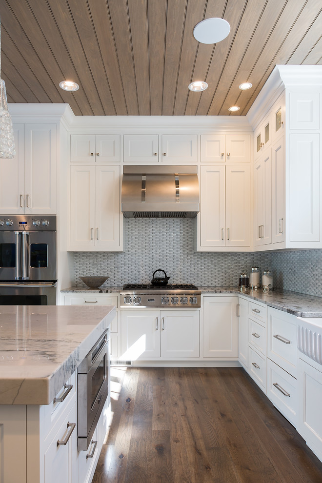 Inspiration for a large transitional l-shaped medium tone wood floor eat-in kitchen remodel with a farmhouse sink, recessed-panel cabinets, white cabinets, marble countertops, gray backsplash, porcelain backsplash, stainless steel appliances and an island