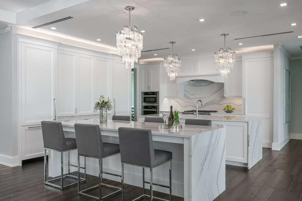 Photo of a beach style kitchen in Tampa with recessed-panel cabinets, white cabinets, marble benchtops, white splashback, marble splashback, stainless steel appliances, dark hardwood floors, multiple islands and white benchtop.