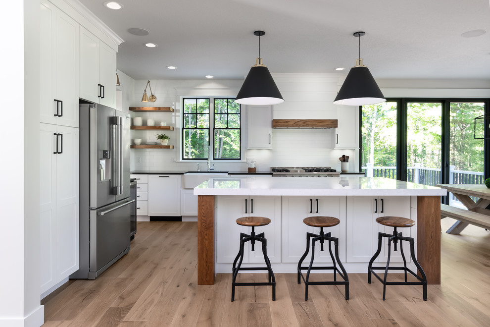 Photo of a country l-shaped kitchen in Other with a farmhouse sink, shaker cabinets, white cabinets, white splashback, subway tile splashback, stainless steel appliances, medium hardwood floors, with island, beige floor and white benchtop.