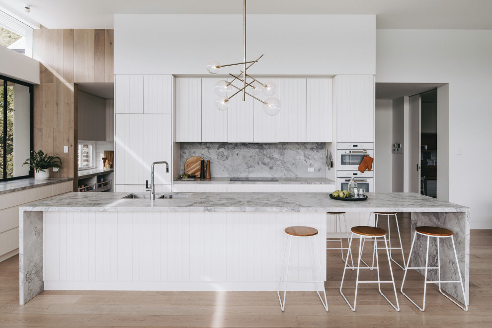 This is an example of a beach style kitchen in Adelaide with an undermount sink, recessed-panel cabinets, marble benchtops, grey splashback, marble splashback, white appliances, medium hardwood floors, a peninsula and grey benchtop.