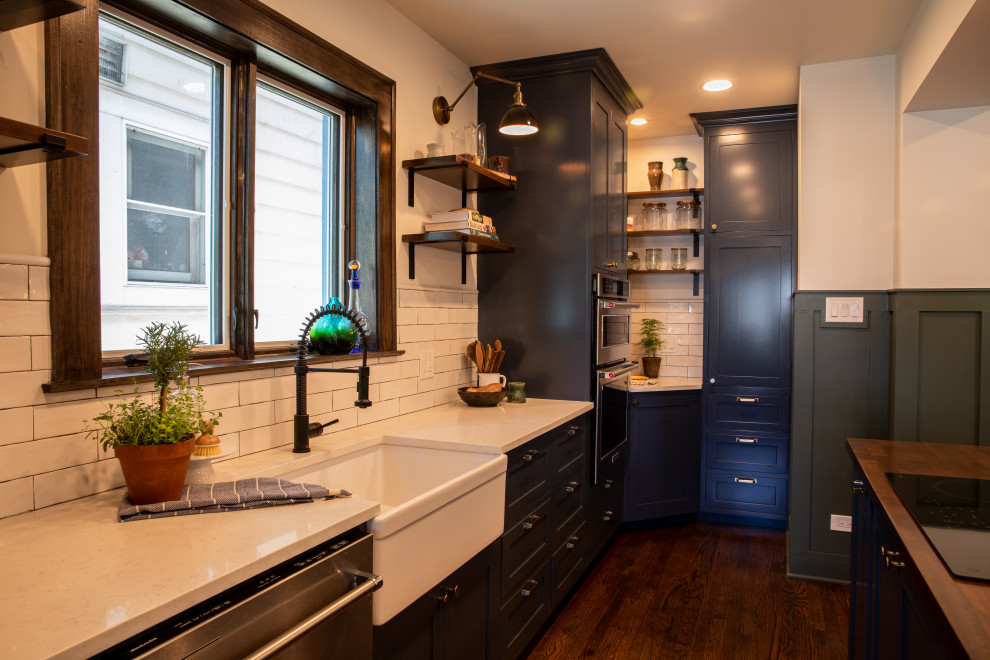 Photo of a medium sized traditional kitchen/diner in Chicago with a belfast sink, shaker cabinets, blue cabinets, engineered stone countertops, white splashback, metro tiled splashback, stainless steel appliances, dark hardwood flooring, an island and white worktops.