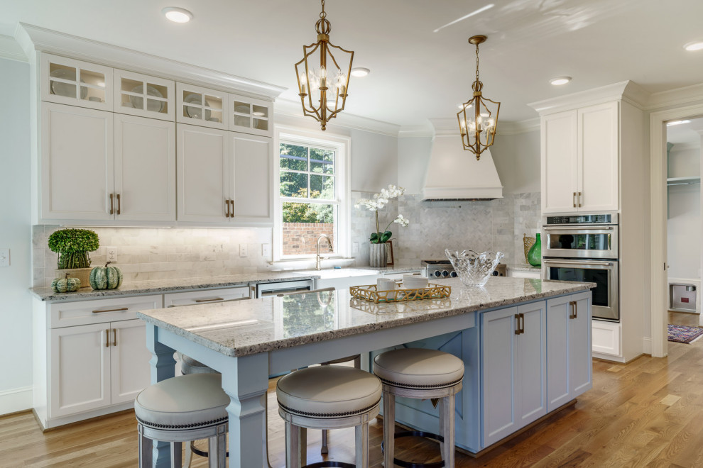 Example of a transitional medium tone wood floor and brown floor kitchen design with a farmhouse sink, shaker cabinets, white cabinets, gray backsplash, stainless steel appliances, an island and gray countertops
