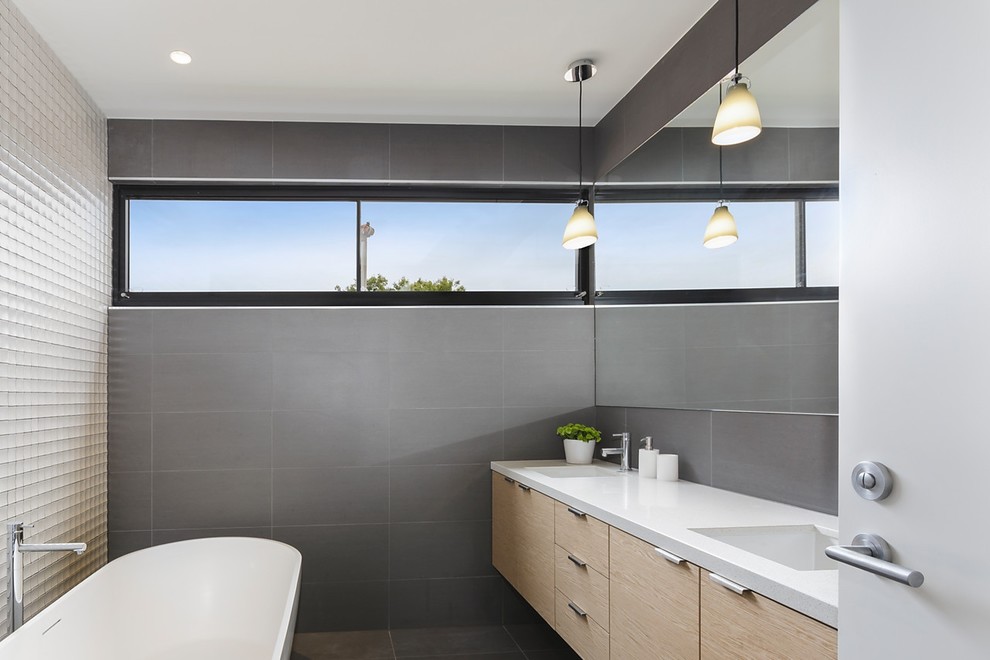 This is an example of a mid-sized modern master bathroom in Melbourne with medium wood cabinets, a freestanding tub, gray tile, ceramic tile, grey walls, ceramic floors, an undermount sink, engineered quartz benchtops, grey floor and flat-panel cabinets.