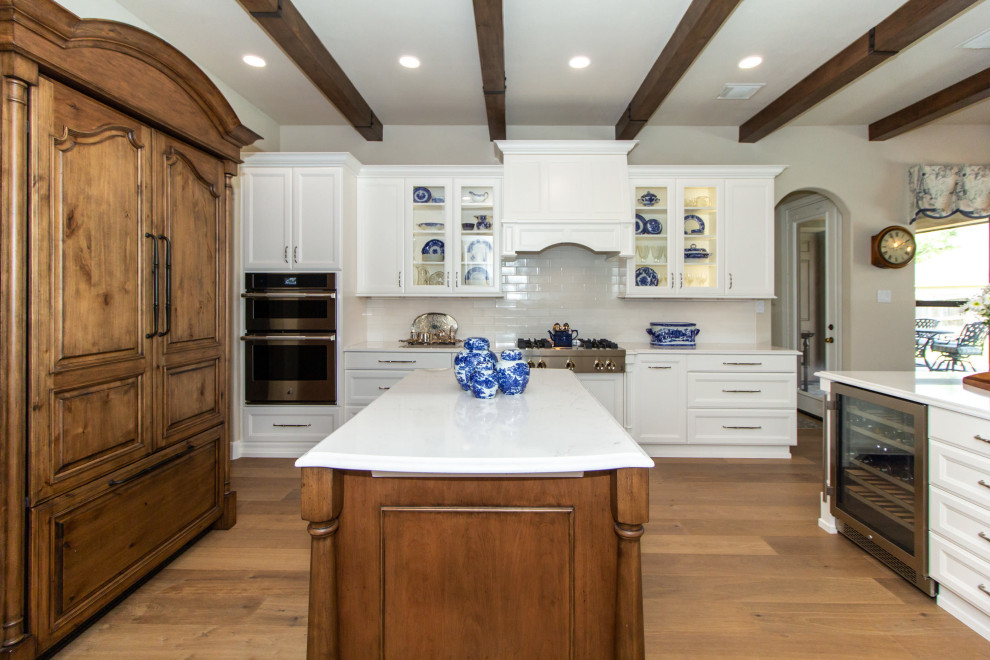 Large kitchen in Houston with an undermount sink, recessed-panel cabinets, white cabinets, quartz benchtops, white splashback, ceramic splashback, stainless steel appliances, medium hardwood floors, with island, brown floor, white benchtop and exposed beam.