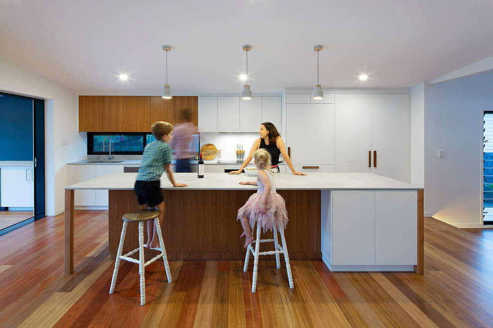 Design ideas for a mid-sized beach style kitchen in Brisbane with an undermount sink, granite benchtops, white splashback, medium hardwood floors, with island, grey benchtop, flat-panel cabinets and white cabinets.