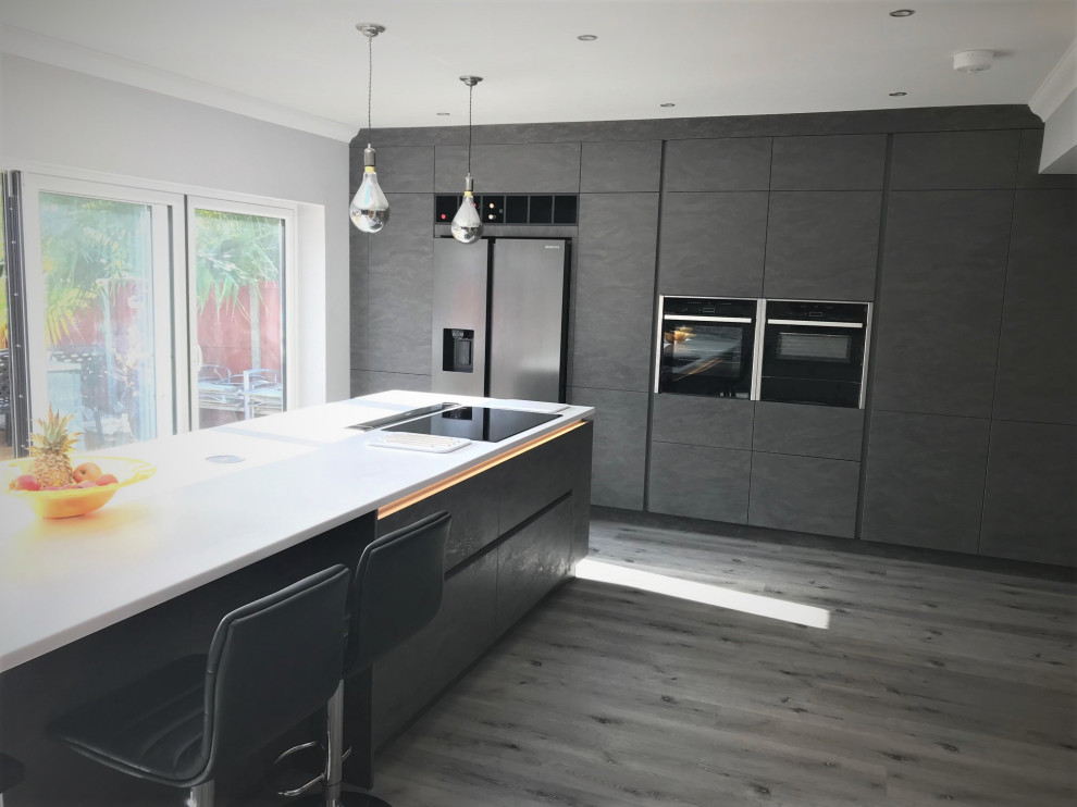 Large modern l-shaped open plan kitchen in Sussex with a drop-in sink, flat-panel cabinets, grey cabinets, solid surface benchtops, orange splashback, glass sheet splashback, black appliances, with island and white benchtop.