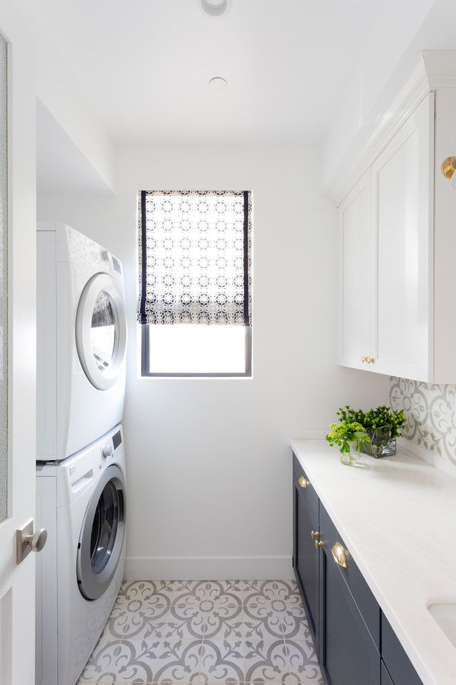 This is an example of a beach style galley dedicated laundry room in Los Angeles with an undermount sink, shaker cabinets, blue cabinets, white walls, a stacked washer and dryer and white benchtop.