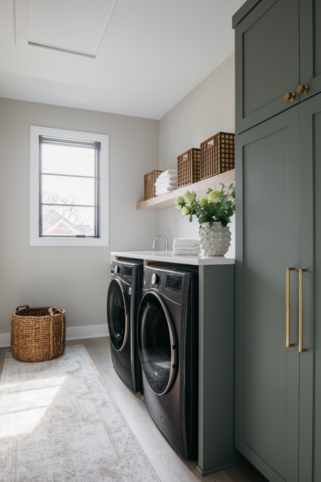 Diseño de cuarto de lavado lineal clásico renovado con armarios estilo shaker, puertas de armario verdes, suelo de madera oscura, lavadora y secadora juntas, suelo marrón y encimeras blancas