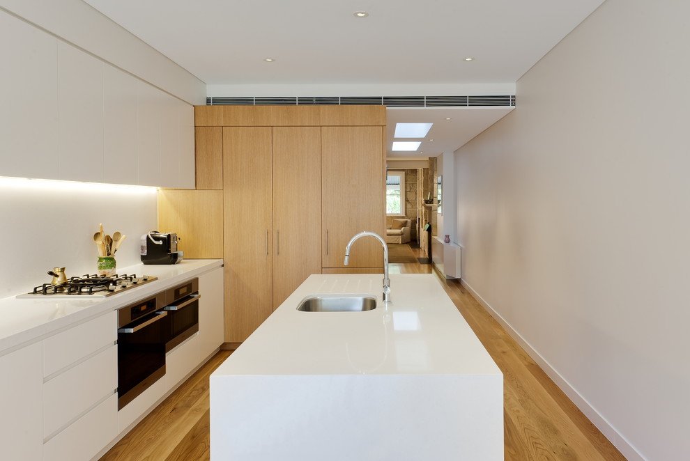 Contemporary l-shaped kitchen in Sydney with white cabinets, solid surface benchtops, white splashback, stainless steel appliances, light hardwood floors, with island, white benchtop, an undermount sink, flat-panel cabinets and brown floor.
