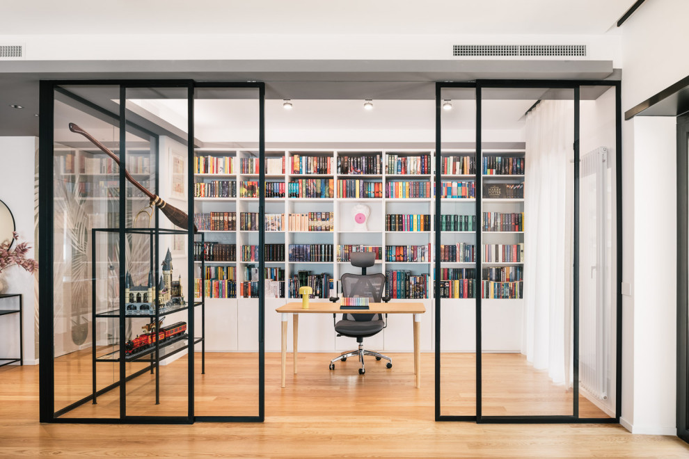 Immagine di un grande studio minimal con libreria, pareti bianche, parquet chiaro, scrivania autoportante e pavimento marrone