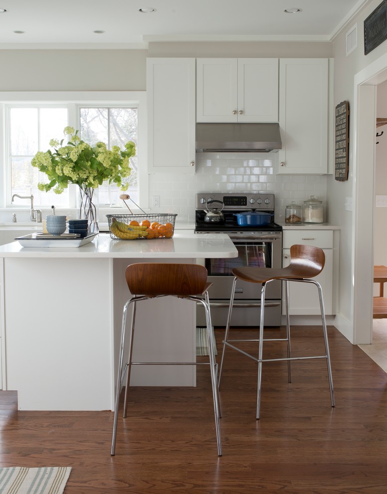 Design ideas for a mid-sized beach style l-shaped eat-in kitchen in New York with shaker cabinets, white cabinets, medium hardwood floors, with island, a farmhouse sink, solid surface benchtops, white splashback, ceramic splashback, stainless steel appliances and brown floor.