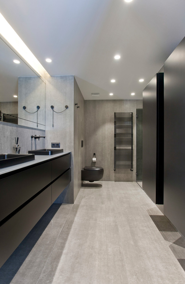 Photo of a mid-sized contemporary powder room in Other with flat-panel cabinets, black cabinets, a wall-mount toilet, gray tile, ceramic tile, grey walls, ceramic floors, a vessel sink, glass benchtops, grey floor and grey benchtops.