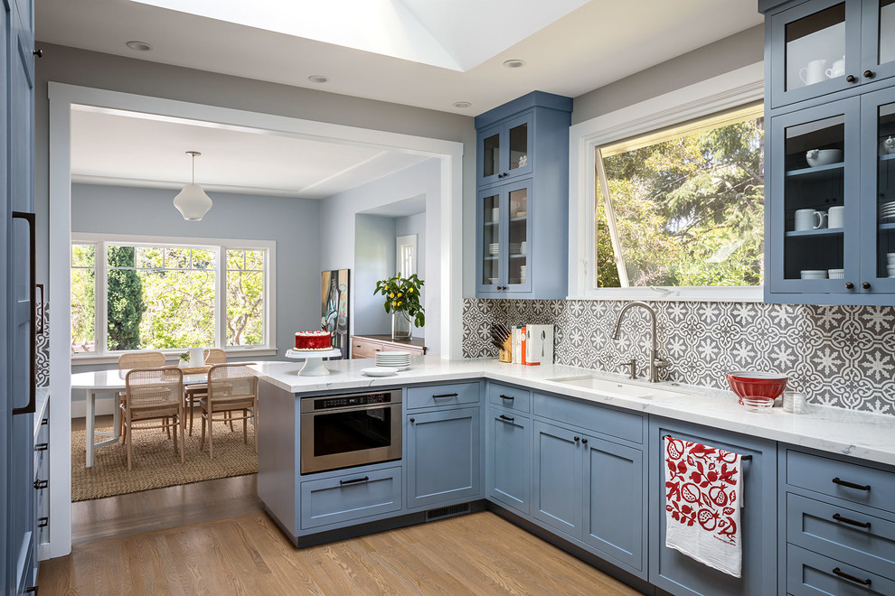 Design ideas for a transitional l-shaped eat-in kitchen in San Francisco with shaker cabinets, blue cabinets, quartz benchtops, cement tile splashback, white benchtop, an undermount sink, multi-coloured splashback, stainless steel appliances, light hardwood floors, a peninsula and beige floor.