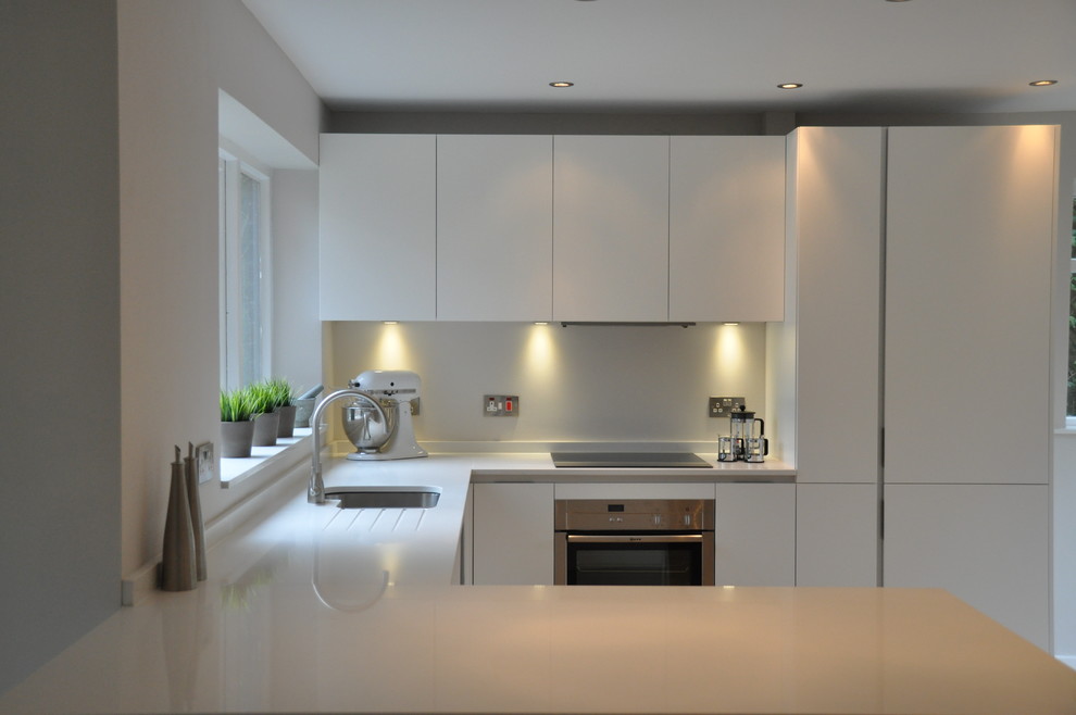 This is an example of a small contemporary u-shaped eat-in kitchen in Cheshire with a drop-in sink, flat-panel cabinets, white cabinets, solid surface benchtops, white splashback, glass sheet splashback, stainless steel appliances, medium hardwood floors and a peninsula.