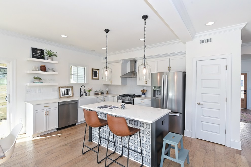 This is an example of a country l-shaped kitchen in Charlotte with a farmhouse sink, shaker cabinets, white cabinets, white splashback, stainless steel appliances, medium hardwood floors, with island, brown floor and white benchtop.