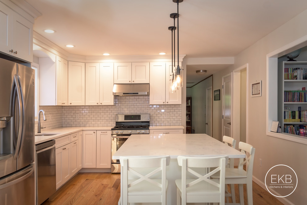 Photo of a mid-sized traditional l-shaped eat-in kitchen in New York with an undermount sink, shaker cabinets, white cabinets, quartz benchtops, white splashback, ceramic splashback, stainless steel appliances, light hardwood floors, with island, brown floor and white benchtop.