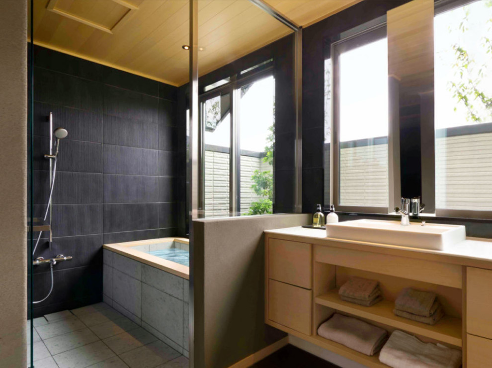 Inredning av ett badrum, med skåp i ljust trä, svart kakel, ett fristående handfat, ett japanskt badkar och våtrum