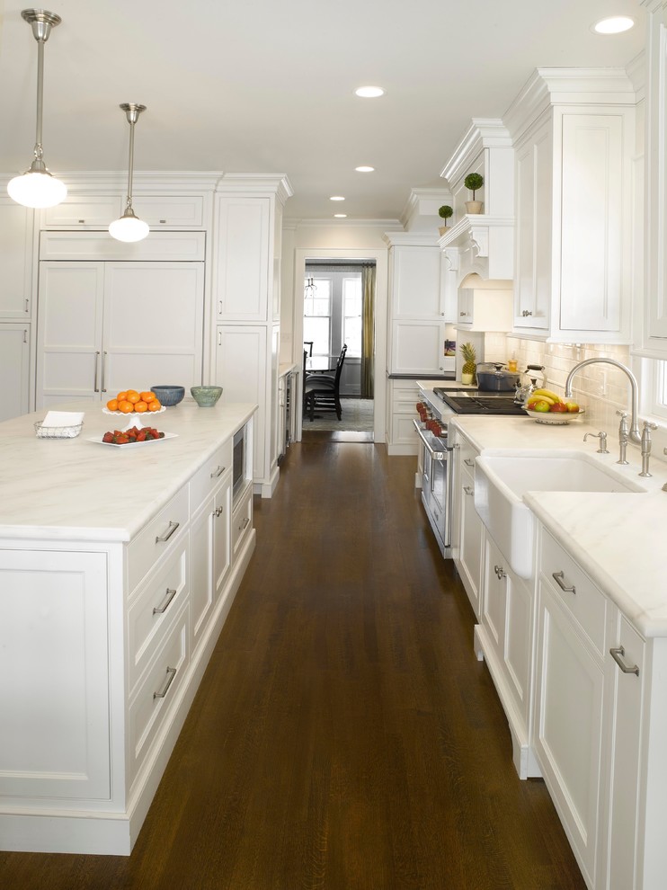 Photo of a traditional eat-in kitchen in New York with a farmhouse sink, white cabinets, marble benchtops, white splashback, subway tile splashback, dark hardwood floors, with island, shaker cabinets and panelled appliances.