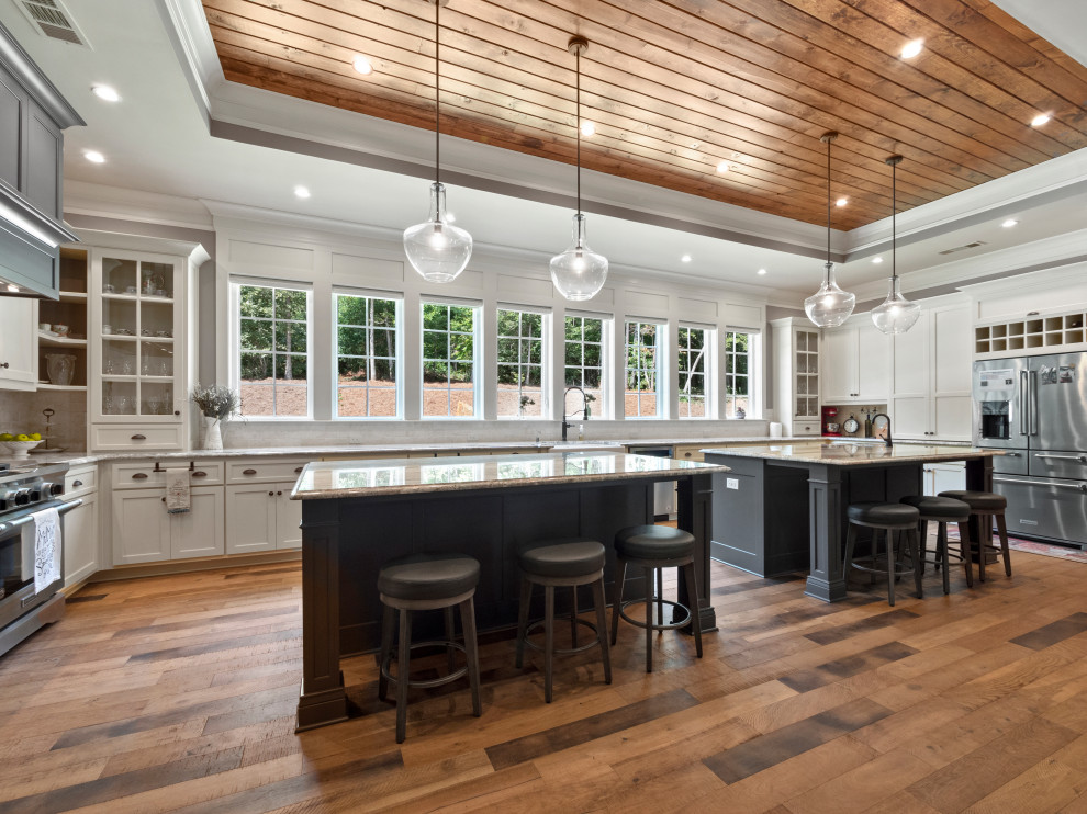 Inspiration for a large country u-shaped kitchen in Atlanta with white cabinets, granite benchtops, stainless steel appliances, medium hardwood floors, multiple islands, brown floor, a farmhouse sink, white splashback, stone tile splashback, grey benchtop and shaker cabinets.