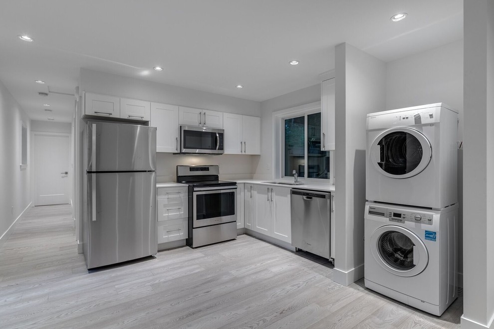Expansive modern l-shaped dedicated laundry room in Vancouver with an undermount sink, shaker cabinets, white cabinets, quartz benchtops, white walls, light hardwood floors, a stacked washer and dryer, grey floor and white benchtop.