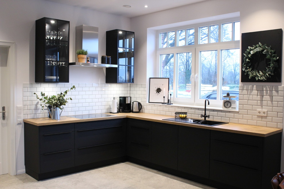 Photo of a mid-sized industrial open plan kitchen in Bremen with a drop-in sink, flat-panel cabinets, black cabinets, white splashback, ceramic splashback, black appliances, slate floors, grey floor and brown benchtop.