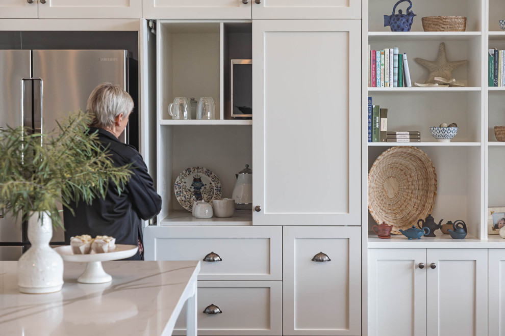 Photo of a large beach style l-shaped open plan kitchen in Adelaide with an undermount sink, shaker cabinets, white cabinets, quartz benchtops, metallic splashback, ceramic splashback, black appliances, laminate floors, with island, brown floor, white benchtop and vaulted.