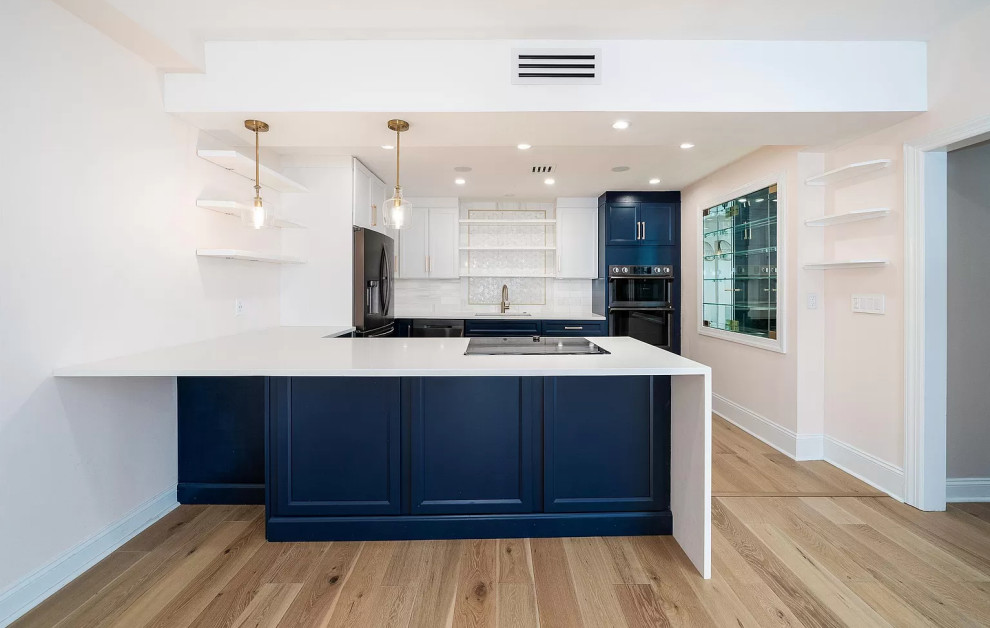 Mid-sized transitional u-shaped eat-in kitchen in New York with an undermount sink, shaker cabinets, blue cabinets, quartz benchtops, white splashback, marble splashback, black appliances, laminate floors, a peninsula, beige floor and white benchtop.
