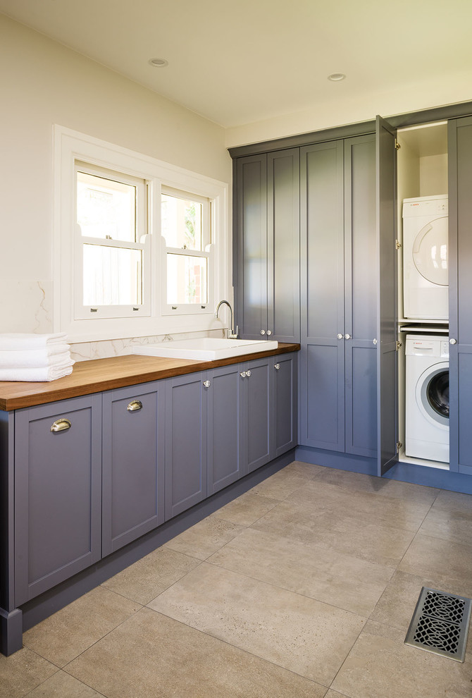 Inspiration for a transitional l-shaped dedicated laundry room in Melbourne with a drop-in sink, shaker cabinets, wood benchtops, beige walls, a stacked washer and dryer, beige floor and beige benchtop.