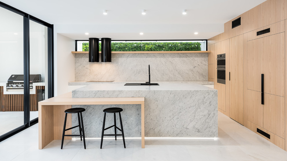 Large modern l-shaped kitchen in Brisbane with light wood cabinets, marble benchtops, marble splashback, black appliances, marble floors, with island, white floor, a drop-in sink, flat-panel cabinets, grey splashback and grey benchtop.