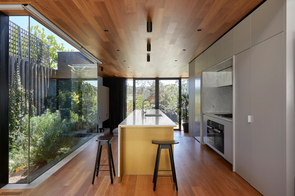 Photo of a mid-sized contemporary galley open plan kitchen in Melbourne with medium hardwood floors, timber, an undermount sink, grey cabinets, marble benchtops, marble splashback, black appliances, with island and white benchtop.