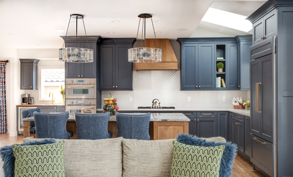Пример оригинального дизайна: большая угловая кухня-гостиная в стиле неоклассика (современная классика) с врезной мойкой, фасадами с утопленной филенкой, синими фасадами, столешницей из кварцита, белым фартуком, фартуком из керамической плитки, техникой под мебельный фасад, светлым паркетным полом, островом, коричневым полом и синей столешницей