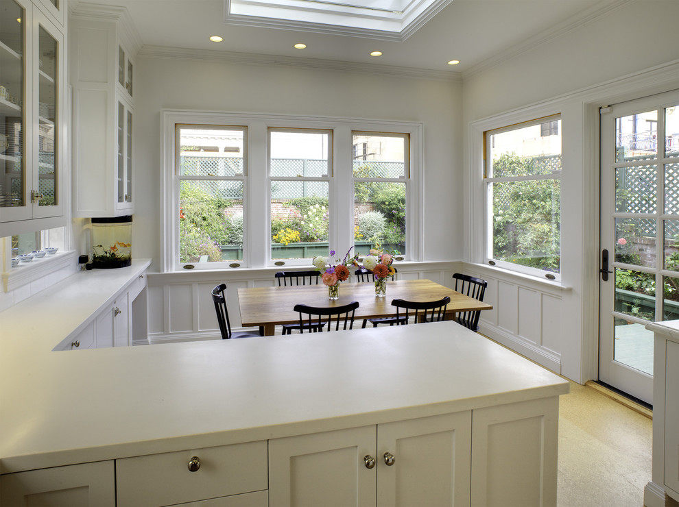 Inspiration for a mid-sized traditional l-shaped eat-in kitchen in San Francisco with glass-front cabinets, white cabinets, solid surface benchtops, white splashback, subway tile splashback, light hardwood floors, a drop-in sink, stainless steel appliances and with island.