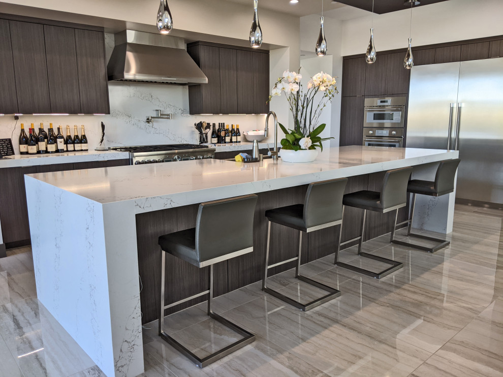 This is an example of an expansive modern l-shaped open plan kitchen in Las Vegas with a belfast sink, flat-panel cabinets, dark wood cabinets, engineered stone countertops, white splashback, engineered quartz splashback, stainless steel appliances, porcelain flooring, an island, beige floors and white worktops.