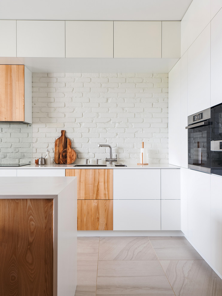 Contemporary l-shaped eat-in kitchen in Other with an undermount sink, flat-panel cabinets, white cabinets, white splashback, brick splashback, stainless steel appliances, with island, beige floor and white benchtop.