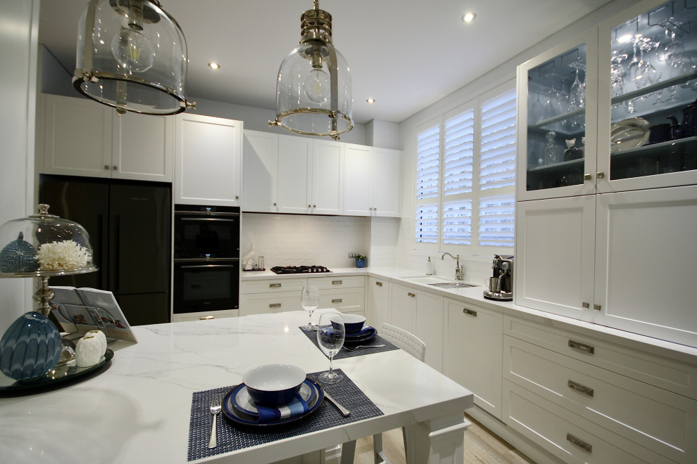 Mid-sized transitional l-shaped open plan kitchen in Sydney with an undermount sink, shaker cabinets, white cabinets, quartz benchtops, white splashback, subway tile splashback, stainless steel appliances, light hardwood floors, with island, brown floor and white benchtop.