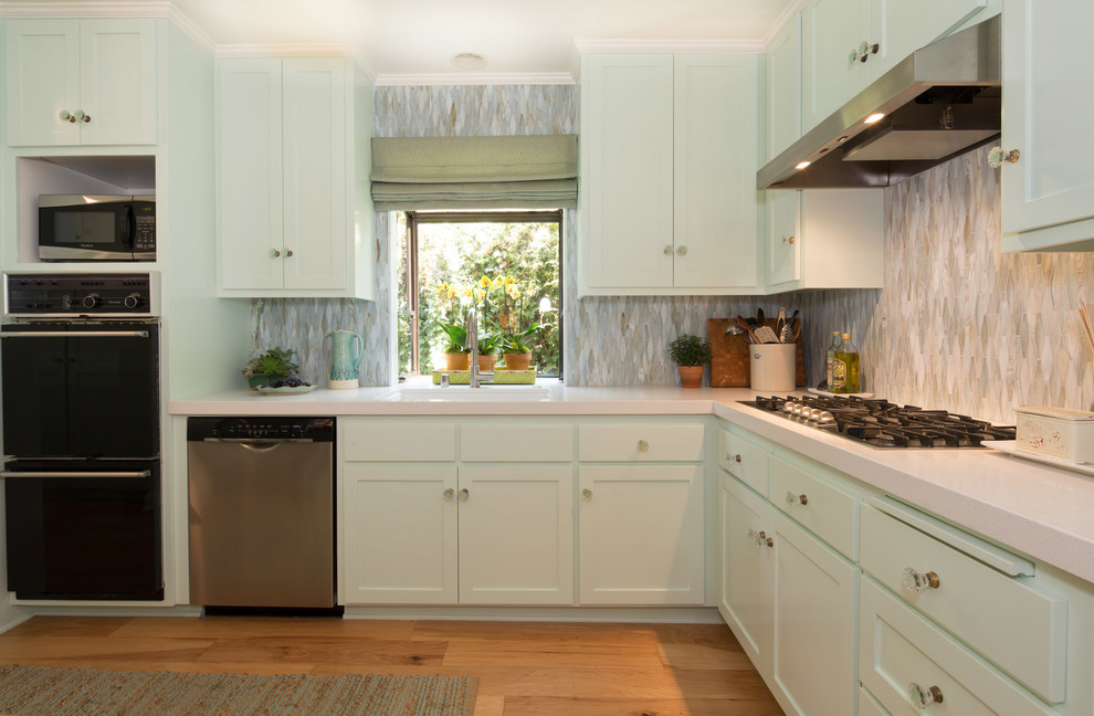 Inspiration for a mid-sized beach style l-shaped separate kitchen in Los Angeles with a peninsula, shaker cabinets, green cabinets, solid surface benchtops, green splashback, glass sheet splashback, stainless steel appliances, a single-bowl sink and medium hardwood floors.