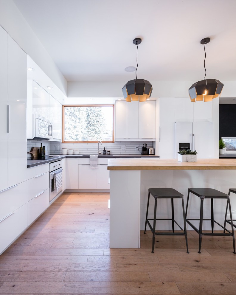 Inspiration for a modern l-shaped kitchen in Los Angeles with an undermount sink, flat-panel cabinets, white cabinets, white splashback, white appliances, medium hardwood floors, with island, brown floor and black benchtop.