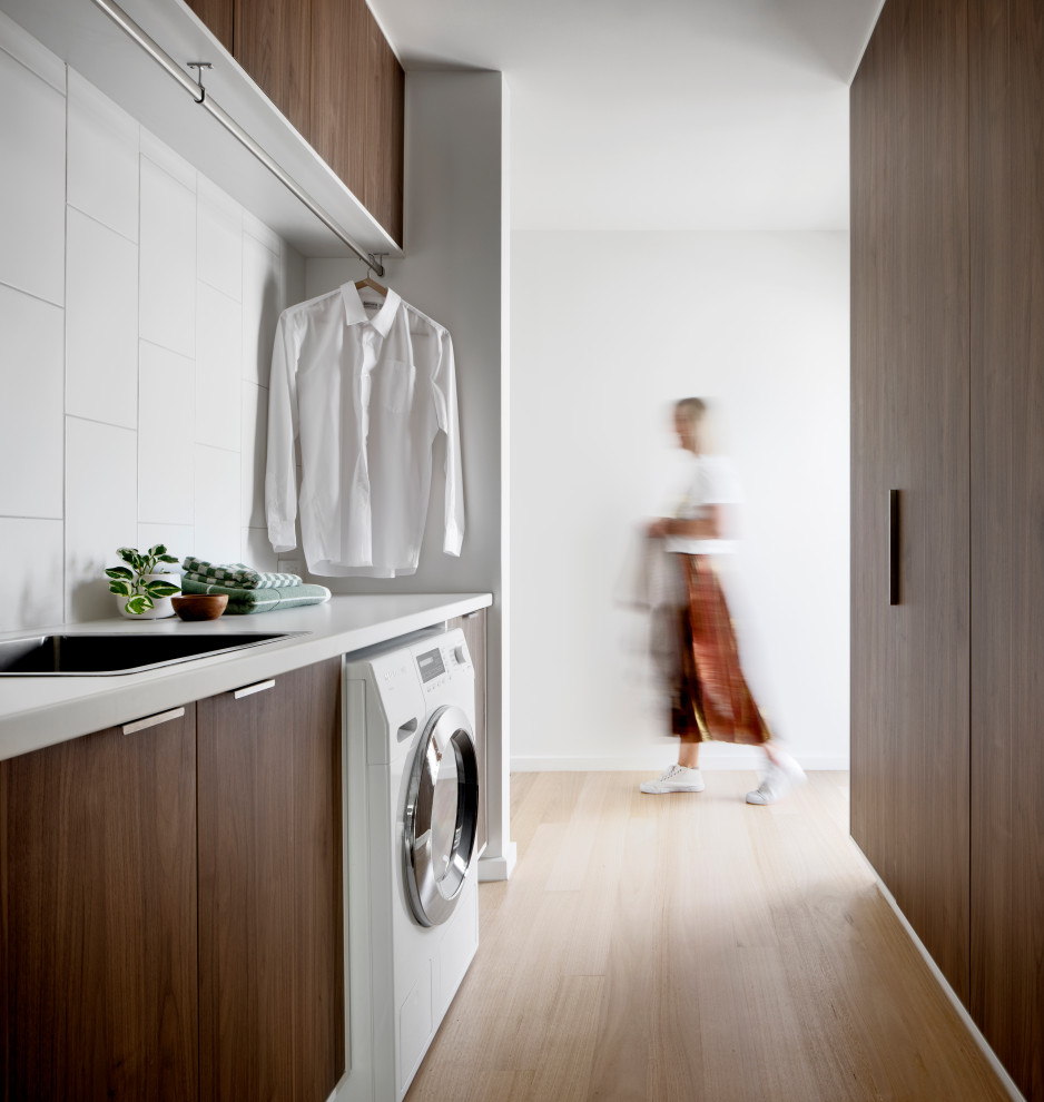 Inspiration for a contemporary galley dedicated laundry room in Melbourne with a drop-in sink, flat-panel cabinets, dark wood cabinets, white splashback, white walls, light hardwood floors and white benchtop.