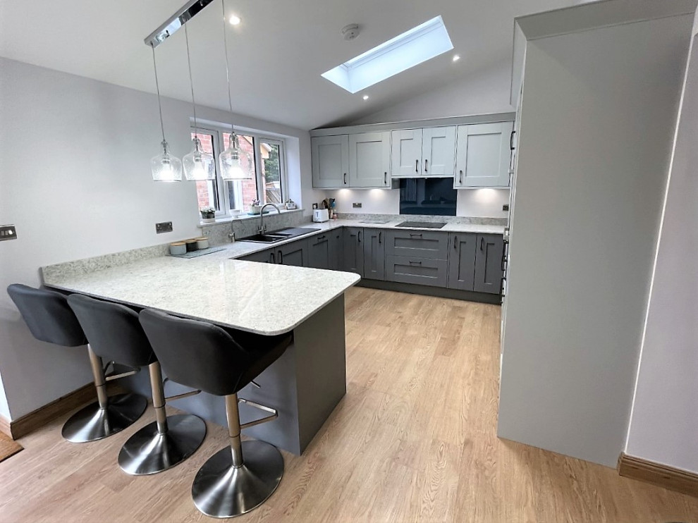 Photo of a mid-sized modern u-shaped eat-in kitchen in Other with a drop-in sink, shaker cabinets, grey cabinets, quartzite benchtops, grey splashback, glass sheet splashback, black appliances, laminate floors, a peninsula, brown floor and white benchtop.