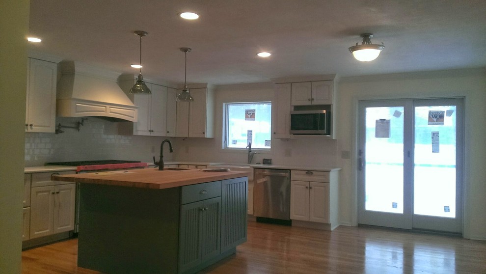 Inspiration for a transitional l-shaped medium tone wood floor kitchen remodel with a farmhouse sink, raised-panel cabinets, white cabinets, white backsplash, subway tile backsplash, stainless steel appliances and an island